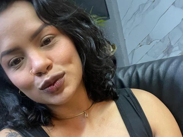 MariGonz, con cabello negro rizado, aro en la nariz, piercing en el labio, collar de oro, camiseta negra sin mangas, labios fruncidos y un tatuaje en el hombro izquierdo, sentada en un sofá de cuero negro..