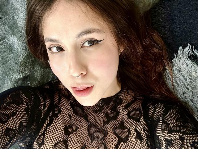 AmberlStha has pale skin, brown eyes, and a nose ring. She's wearing a black lace top, with her brown hair spread out. Her lips are slightly parted.