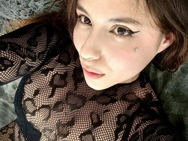 AmberlStha with fair skin, brown eyes, and a nose ring, wearing a black lace top, showing her breasts through the mesh, with a small diamond stud on her left cheek.