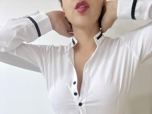 AriaPritzker with hands behind head, wearing white button-up shirt, black stripes on sleeves, pink lipstick, showing cleavage, shirt slightly open, white background.