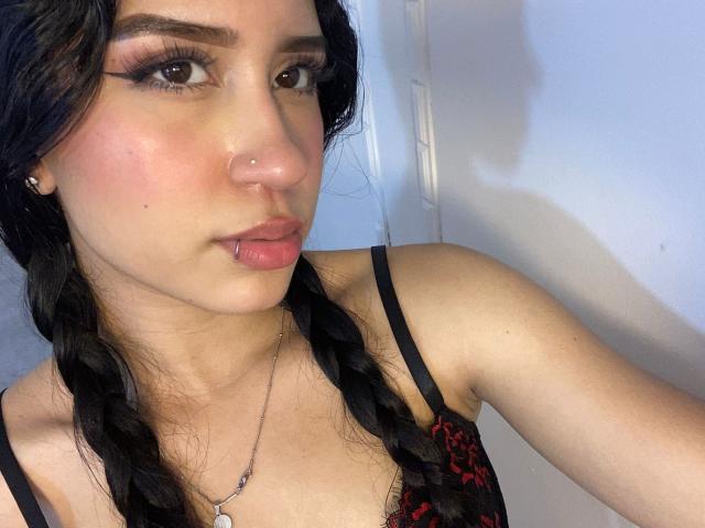 AnnaTorress, con cabello negro trenzado, aro en la nariz y piercing en el labio, vestida con una blusa de encaje negra y roja, mostrando sus labios rosados y sus cejas oscuras y pobladas..