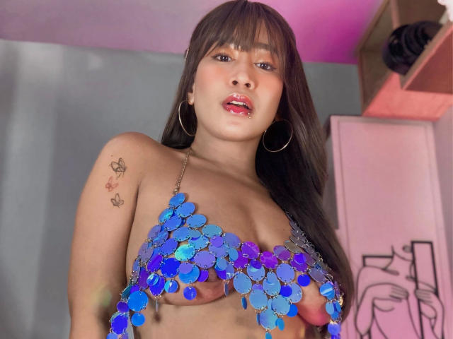 OHCUMTOMYTITS-hot with long brown hair, wearing a blue sequin bra covering her tits, hoop earrings, tattoo on right shoulder, pink lipstick, and a slight open-mouthed expression.