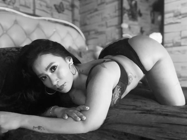 OHCUMTOMYTITS-hot is on all fours on a bed, wearing black lace panties, with a tattoo on her arm and hoop earrings, in a black-and-white photo.