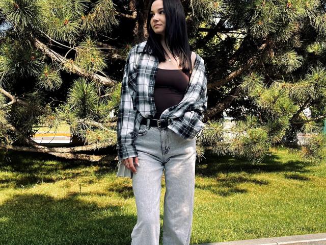 Treat-hot with black hair, wearing a black tank top, plaid shirt, and light blue jeans, standing in front of a pine tree, showing her ass with her hands down.