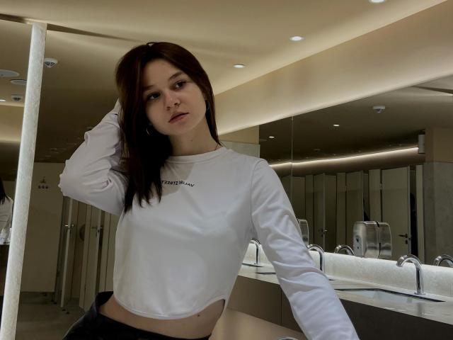 Treat-hot with long brown hair, white crop top, and black pants, standing in a bathroom, hand in hair, mirror reflecting background, slightly parted lips, neutral expression.