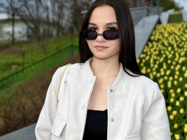 Mulher com cabelos castanhos longos, usando óculos de sol pretos, uma jaqueta branca e uma blusa preta, em pé na frente de flores amarelas, sorrindo discretamente. Suas nádegas não estão visíveis..