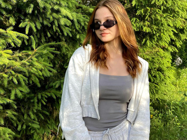 Treat-hot with long brown hair, wearing black sunglasses, a gray tank top, and a white zip-up, standing in front of green bushes, looking slightly to the side.