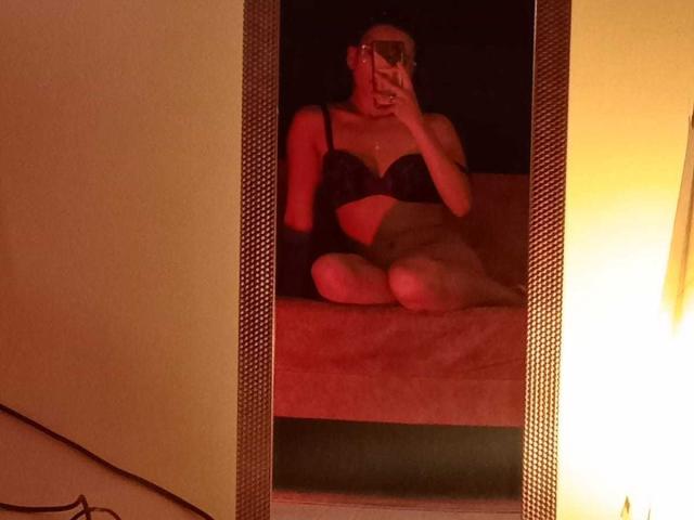 KateDonovan zit voor een spiegel, gekleed in zwarte lingerie waarbij haar borsten en schaamstreek zichtbaar zijn, en maakt een selfie met haar telefoon, met gekruiste benen, in gedimd rood licht..