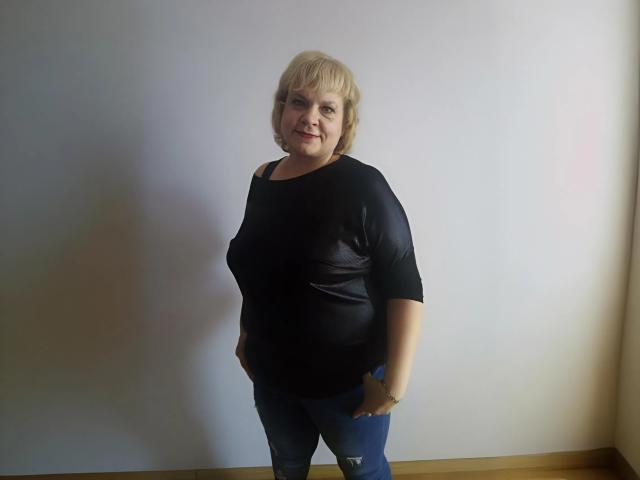 BeverlyCynia is a chubby woman with short blonde hair, wearing a black shirt and blue jeans, standing with hands in pockets, smiling at the camera.