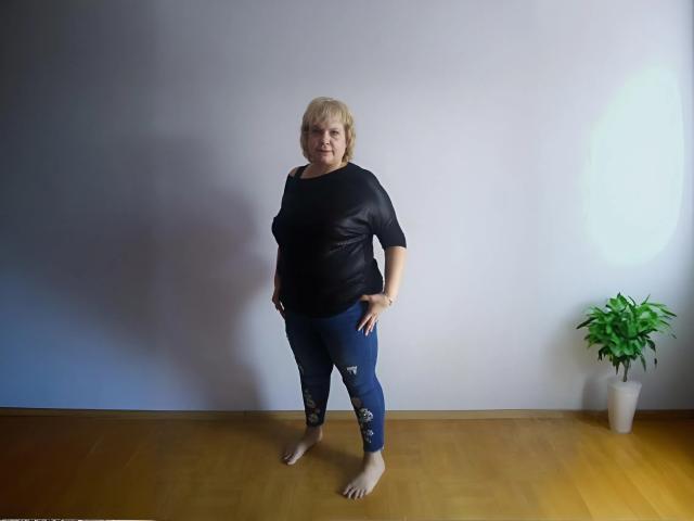 BeverlyCynia stands barefoot in a room, wearing a black shirt, blue ripped pants, and a bracelet, with a potted green plant to the right.