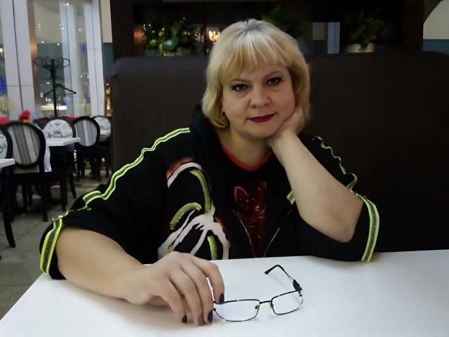 BeverlyCynia with blonde hair, red lipstick, black and neon-green shirt, resting head on hand, wearing glasses on white table, sitting in restaurant. Her tits are small.