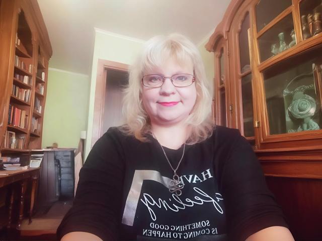 BeverlyCynia, a chubby woman with blonde hair and glasses, wearing a black shirt, smiling at the camera with pink lipstick, has a necklace and is in a wooden room.