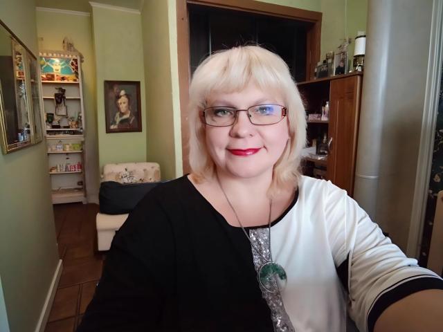 BeverlyCynia, blonde with glasses, red lipstick, black and white shirt, green pendant, smiling, fair skin, average tits, medium build, standing in a room, taking a selfie.