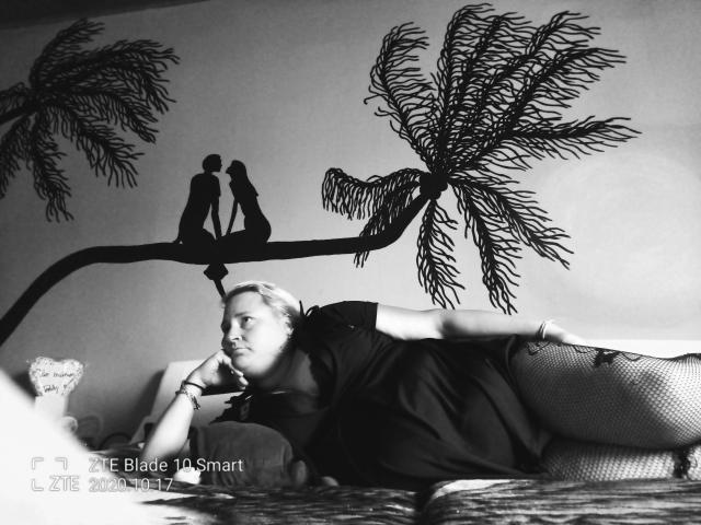 KleinerTeufel82-hot is lying on a couch, wearing a dark shirt and fishnet pants, with a pensive expression. Black palm tree wall decal with silhouetted couple above.