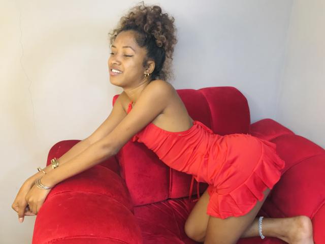 SaynahSisi with curly brown hair, smiling, wearing a red ruched dress, kneeling on a red velvet couch, arms resting on the back. Her ass is visible, looking cute.