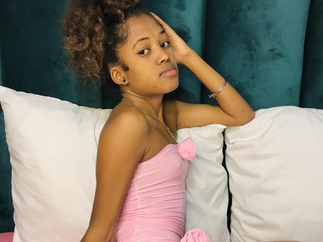 SaynahSisi with curly brown hair, wearing a pink strapless dress with a bow, sitting on white pillows, hand on head, looking to the side, dark green velvet background.