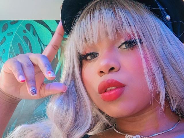 LizzyFoxy with platinum blonde hair, black hat, pink lipstick, blue eyes, and red nails, showing off a purple nail design, looking into the camera.