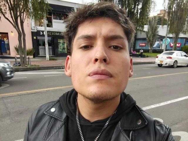 DiegoBiker with short, curly brown hair, light brown skin, and a slight mustache, wearing a black leather jacket and a black hoodie, standing on a street.