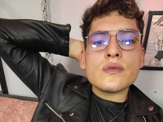 DiegoBiker with curly brown hair, wearing a black leather jacket, glasses with blue reflections, and a black shirt, pouting with his hand behind his head.