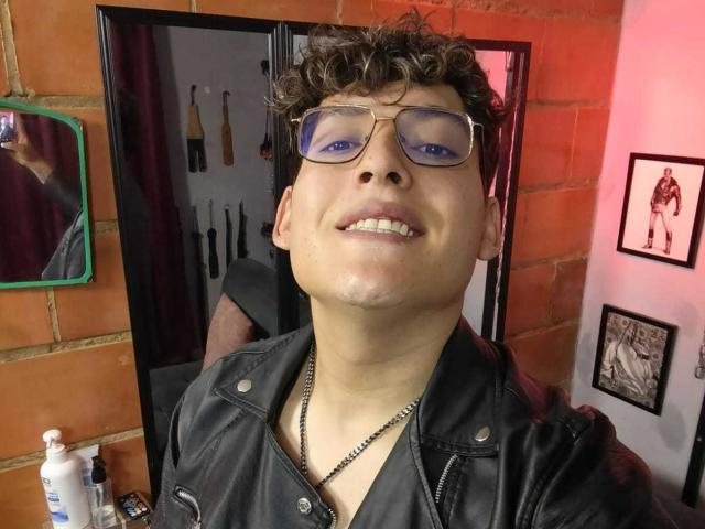 DiegoBiker with curly brown hair, blue eyeshadow, and glasses, wearing a black leather jacket, smiling at the camera, standing in front of a mirror.