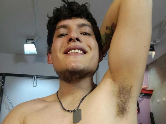 DiegoBiker, shirtless, smiling with arm raised, showing hairy armpit, black curly hair, small beard, tattoo on arm, necklace with dog tag, light skin, brown eyes.