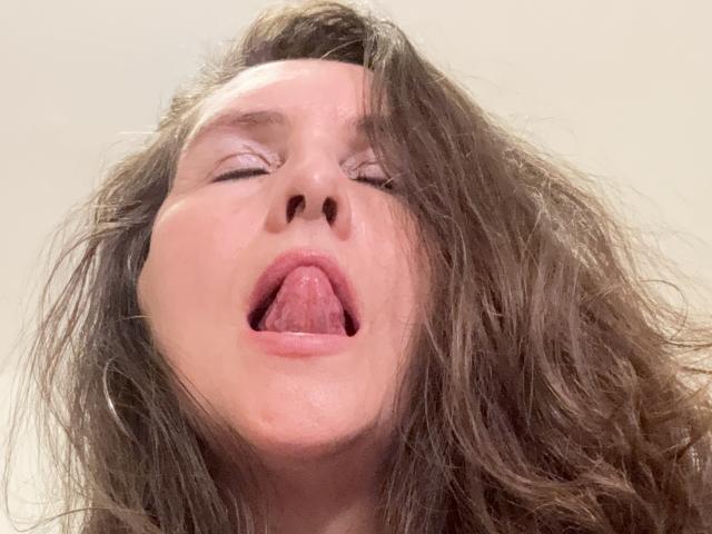 PamelaPalmer's face, eyes closed, tongue out, messy brown hair, slightly flushed, nose up, mouth open, looking somewhat aroused or exhausted.