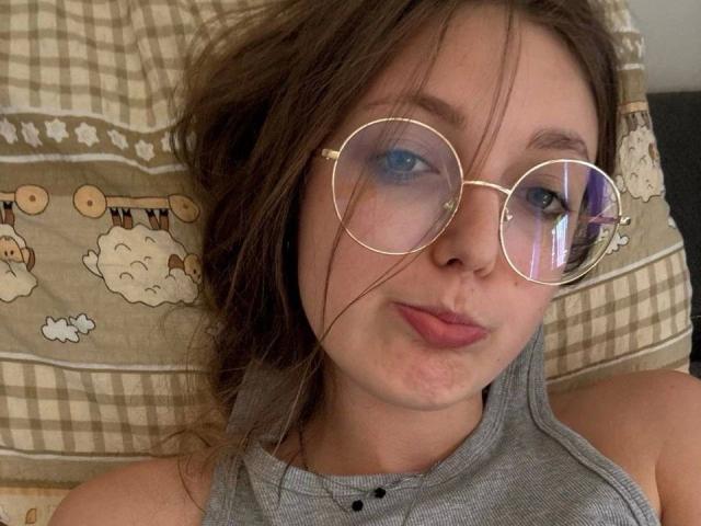 Sara2005-hot a les yeux bleus gonflés, porte des lunettes rondes dorées et a un visage boudeur avec des lèvres légèrement gonflées. Elle porte un haut gris sans manches et a les cheveux bruns en désordre..