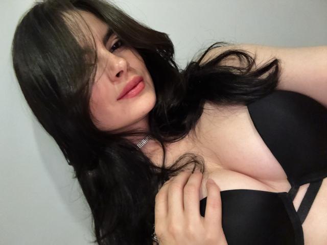 ClariceMontclair with long black hair, pink lips, and black bra, holding her large, shiny tits, wearing a silver necklace, looking seductively at the camera.