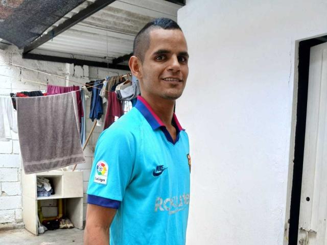 ThiagoDelicius, com cabelo curto e escuro, camisa de futebol azul, gola vermelha, sorrindo, em pé em um quarto simples, toalha pendurada, roupas em um varal, parede branca, prateleira pequena, logotipo esportivo..