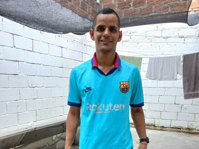 ThiagoDelicius, un homme aux cheveux courts, portant un maillot de foot bleu avec l'inscription "Pekuten" et un écusson de club, souriant, debout dans une ruelle en briques blanches avec des vêtements suspendus..