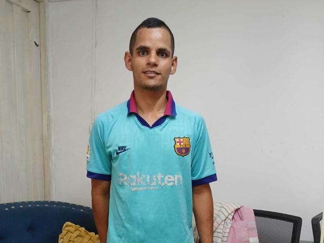 ThiagoDelicius, cabelo preto curto, pele morena, vestindo uma camisa de futebol do FC Barcelona em tons de azul turquesa com gola rosa, em pé em um quarto, com um leve sorriso, braços ao lado do corpo..