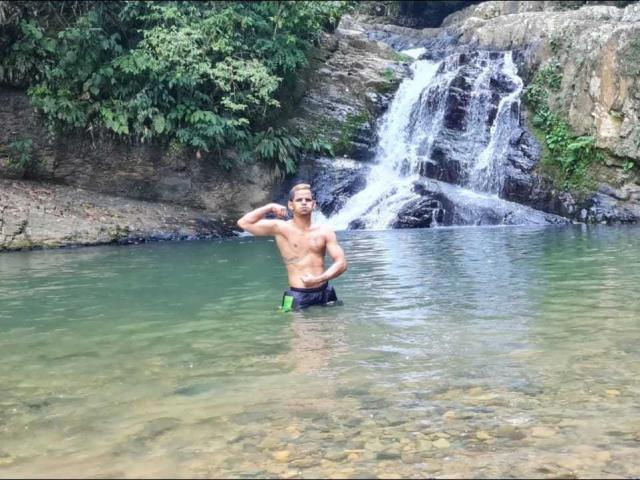 ThiagoDelicius, ohne Hemd, steht in einem felsigen Bach, das Wasser reicht bis zu seinen Oberschenkeln, er trägt grüne und schwarze Badehosen, eine Hand hinter dem Kopf, im Hintergrund ein Wasserfall, er lächelt..