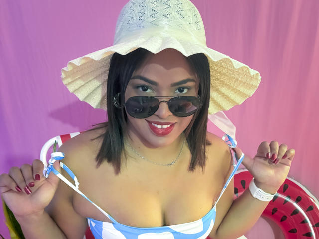 AliceBooty, com seios médios, usando um chapéu de sol branco, um vestido listrado azul e branco, e óculos de sol escuros, sorrindo com batom vermelho, segurando as alças do vestido, fundo rosa..