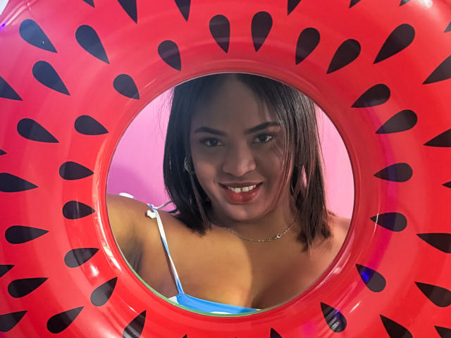 AliceBooty, con senos de tamaño medio, sonriendo a través de un aro inflable rojo con patrones de lágrimas negras, vestida con un top de bikini azul, tiene cabello castaño y un collar de plata..