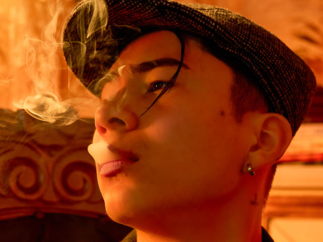 NoahAndRose avec une cigarette, de la fumée sur le visage, portant une casquette plate, des boucles d'oreilles et des cheveux courts, regardant sur le côté, éclairage orange chaud, bouche légèrement ouverte, expression faciale douce..