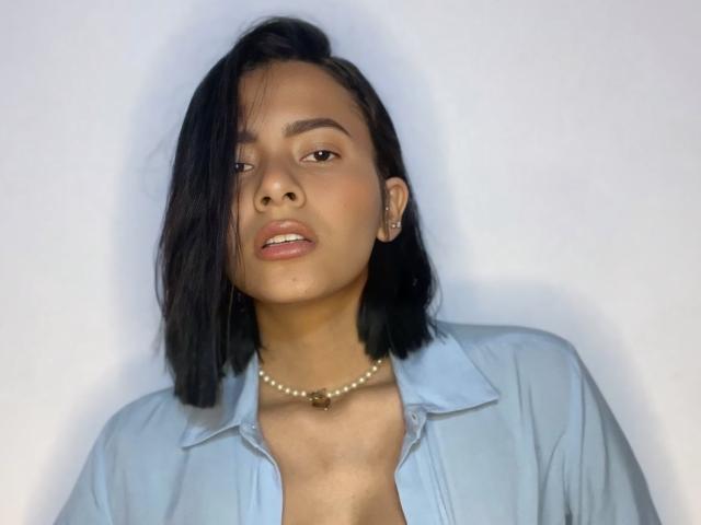 SophieMillerX with black shoulder-length hair, light brown skin, wearing a light blue shirt, necklace, and earrings, looking slightly to the side with parted lips.