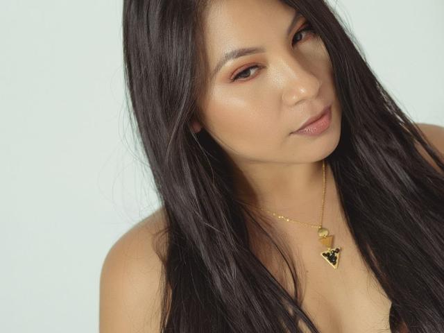 DominikaR with long black hair, brown eyes, and a gold necklace with a black heart pendant, has a slight smile and is looking at the camera.