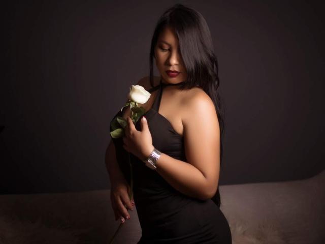 DominikaR with long black hair, holding a white rose, wearing a black halter dress, looking down, her ass slightly visible, a silver watch on her wrist, dark background.