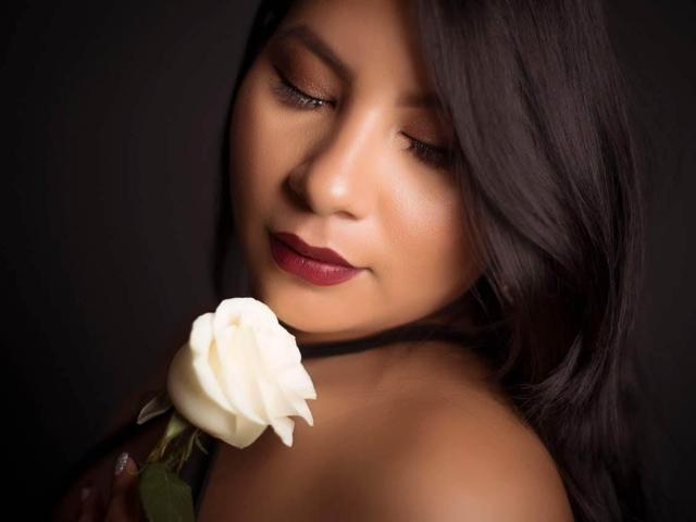 DominikaR with dark hair, closed eyes, and red lipstick, holding a white rose near her nose, her bare shoulders visible. She's looking down at the rose with a gentle smile.