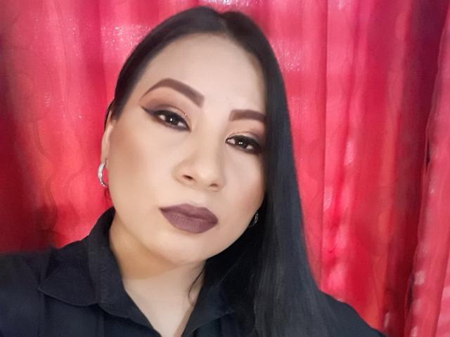 DominikaR has dark, straight hair, thick eyebrows, and is wearing matte brown lipstick with dark eyeliner. She's looking at the camera with a slight pout. Wearing a black shirt.