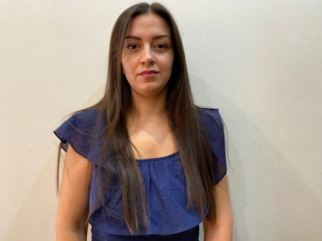 KarinaClover hat langes, braunes Haar, ein ernstes Gesicht und trägt ein blaues Oberteil mit Rüschenärmeln. Ihre Brüste sind klein und ihre Vulva ist nicht sichtbar..