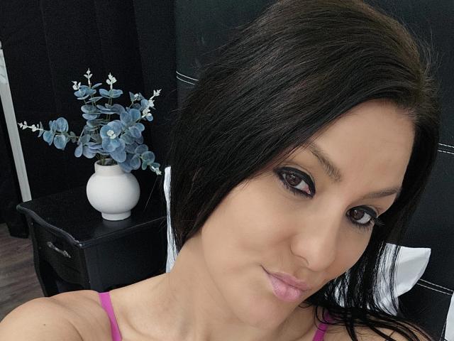 JoliDanielle has dark brown hair, brown eyes, and pink lipstick, wearing a pink top, with a white vase of blue flowers on a black nightstand in the background.