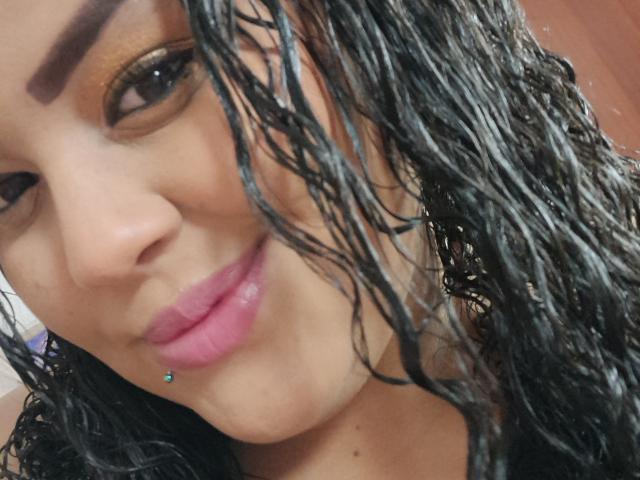 O rosto de Melannie Carter, cabelo cacheado preto e úmido, lábios rosados e brilhantes, pequeno piercing azul nos lábios, sobrancelhas escuras, olhos castanhos, pele clara, sorriso discreto, selfie em close-up, luz natural..