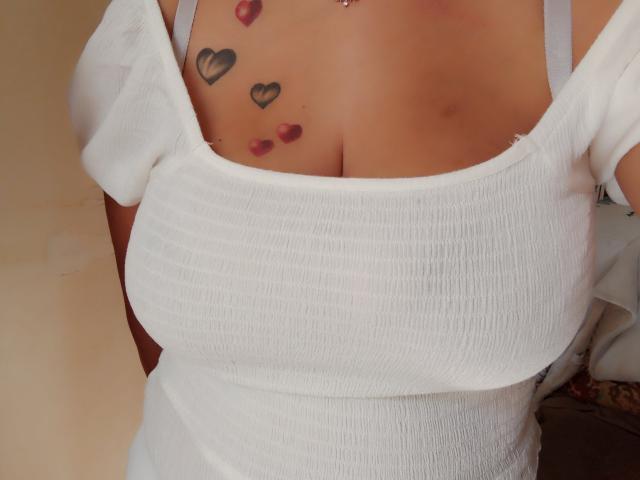 ClemenceFuerte's chest with white ribbed tank top, showing cleavage, has black heart tattoos and red lipstick kisses above the chest. ClemenceFuerte has tan skin and a silver necklace.
