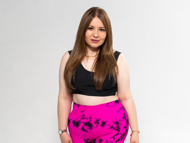 Katte Adams mit langen, braunen Haaren, schwarzem Crop-Top, pinken und schwarzen Hosen mit Muster, kleinen Brüsten, kurvigen Hüften, lächelnd, weißer Hintergrund, trägt eine goldene Halskette und eine Armbanduhr..