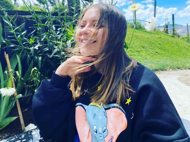 KatteAdams with dirty blonde hair, smiling, wearing a black sweatshirt with a blue cartoon elephant, wind blowing through her hair, standing in a sunny garden.