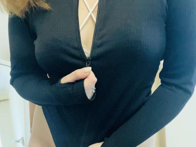KarKutie vestindo uma blusa preta de manga comprida com zíper frontal e amarração na frente, segurando o zíper com a mão direita, expondo os seios, mostrando o bumbum nu..
