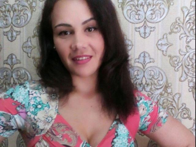 DayanaSins, com cabelos castanho-escuros ondulados, batom rosa e um vestido floral que revela o decote, sorrindo para a câmera, exibindo seus seios..