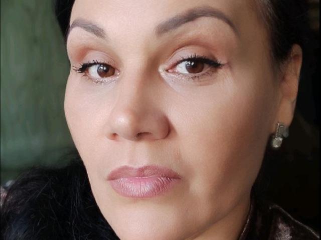 DayanaSins tiene una expresión seria, con ojos marrones, labios rosados y brillantes, y cejas oscuras. Lleva pendientes de diamantes y tiene un tono de piel claro con cabello negro..