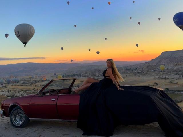 SinnersMuse with long blonde hair, black dress, and bare legs sits on red convertible, surrounded by hot air balloons at sunset. Her dress is billowing, showing her ass.